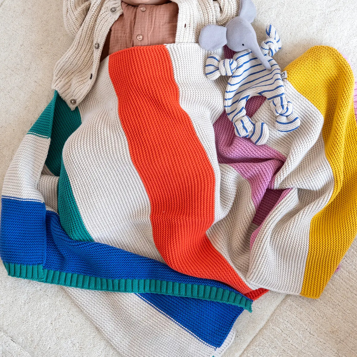 A colorful striped knit blanket with cream, orange, white, teal, blue, and yellow sections, styled with a beige cardigan, a pink shirt, and a blue-and-white striped elephant toy on a cream fabric background.