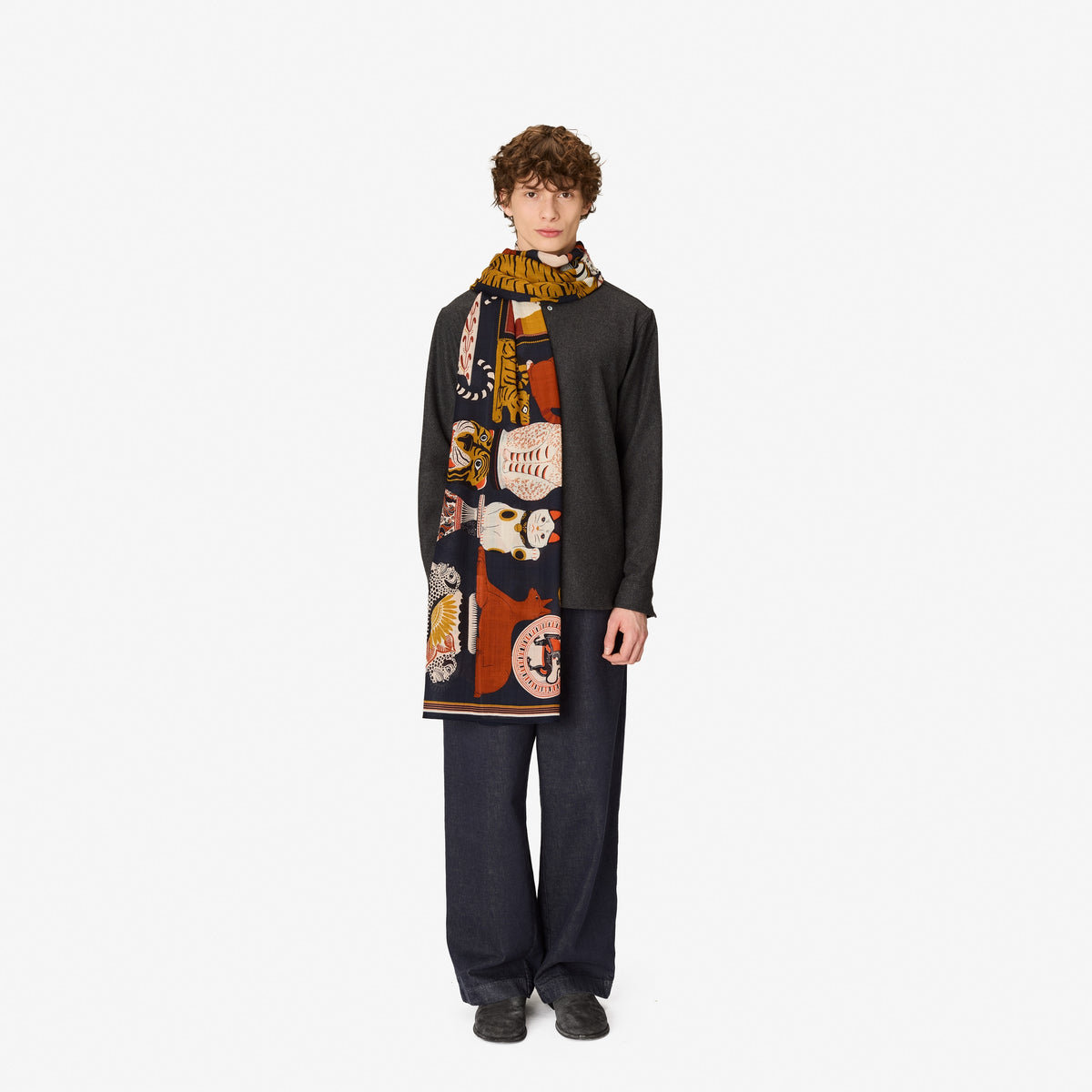 A model stands against a plain white studio background wearing a long black cardigan, dark wide-leg trousers, and a thick patterned scarf with orange, cream, and yellow motifs.