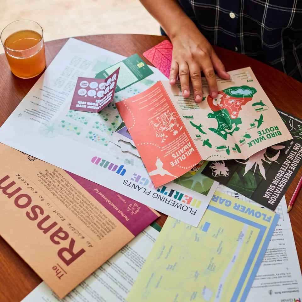 A person's hand rests on a spread of colorful printed cards, booklets, and papers on a round wooden table, with a glass of orange drink nearby.