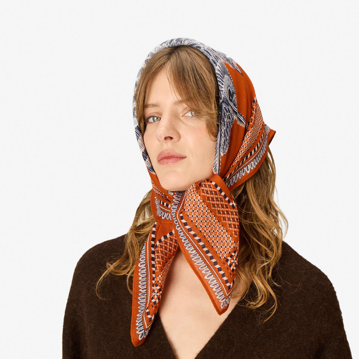 A woman wearing a patterned orange, gray, and white headscarf tied under her head, paired with a brown knit sweater, set against a plain white background.