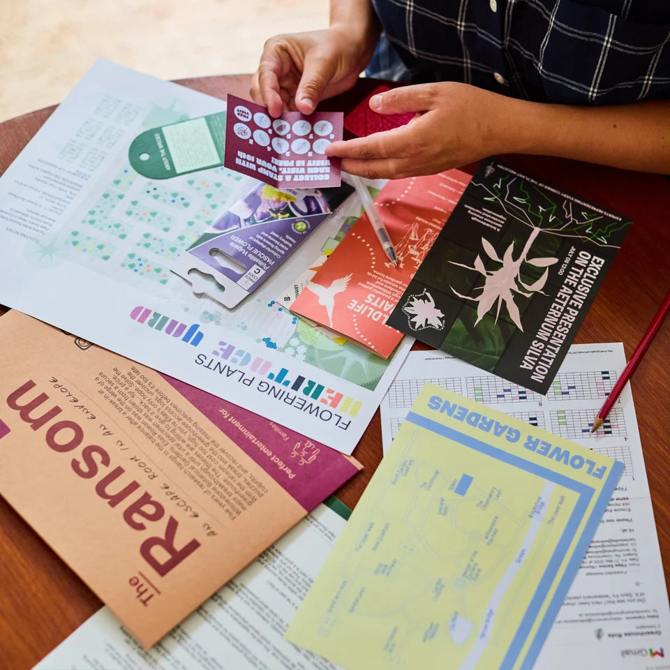 A person's hands holding a small printed card over a spread of gardening and educational printed materials on a wooden table. Visible printed items include a brown booklet titled 'Ransom', a black card reading 'Escape the Dark Night Garden', and other colorful printed sheets.