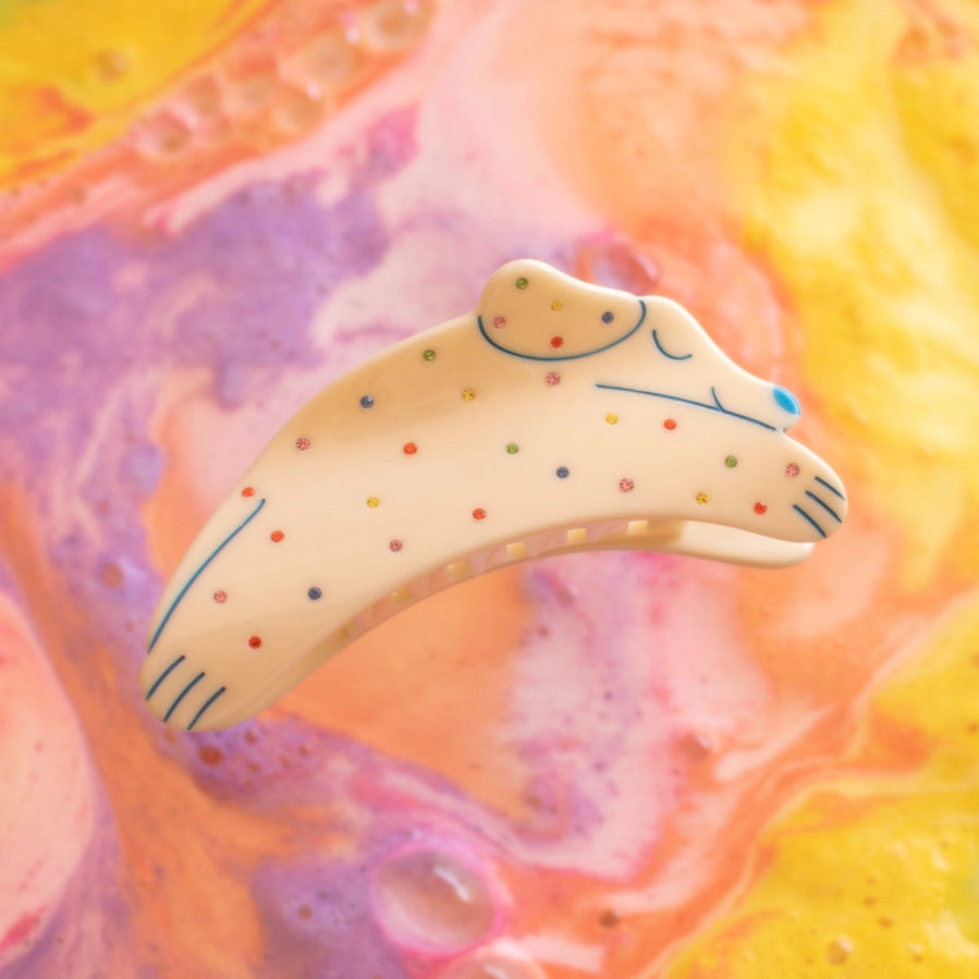 A cream-colored hair claw clip shaped like a cat, decorated with colorful dots and blue line details, set against a vibrant, abstract marbled background of yellow, pink, and purple.