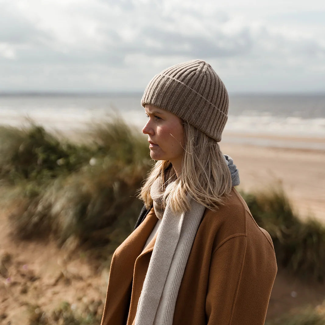 A woman standing on a sandy beach dune wearing a camel brown coat, a cream knit scarf, a taupe ribbed beanie, and matching knit gloves, with the ocean and grassy dunes blurred in the background.