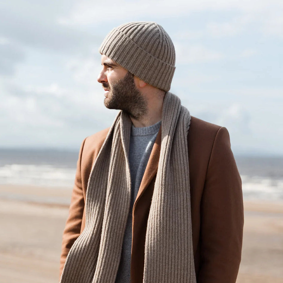 A man standing on a beach dune wearing a brown coat, a thick ribbed taupe scarf, a matching ribbed beanie, a gray t-shirt, and dark jeans, looking off-camera.