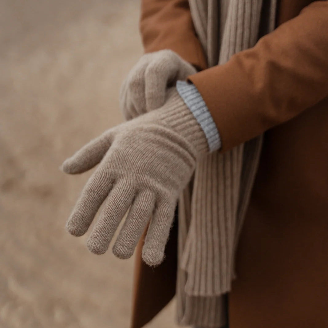 A person outdoors wearing a brown coat and a beige ribbed scarf, with one hand adjusting a beige knit glove. The background is a softly blurred sandy beach or outdoor ground.