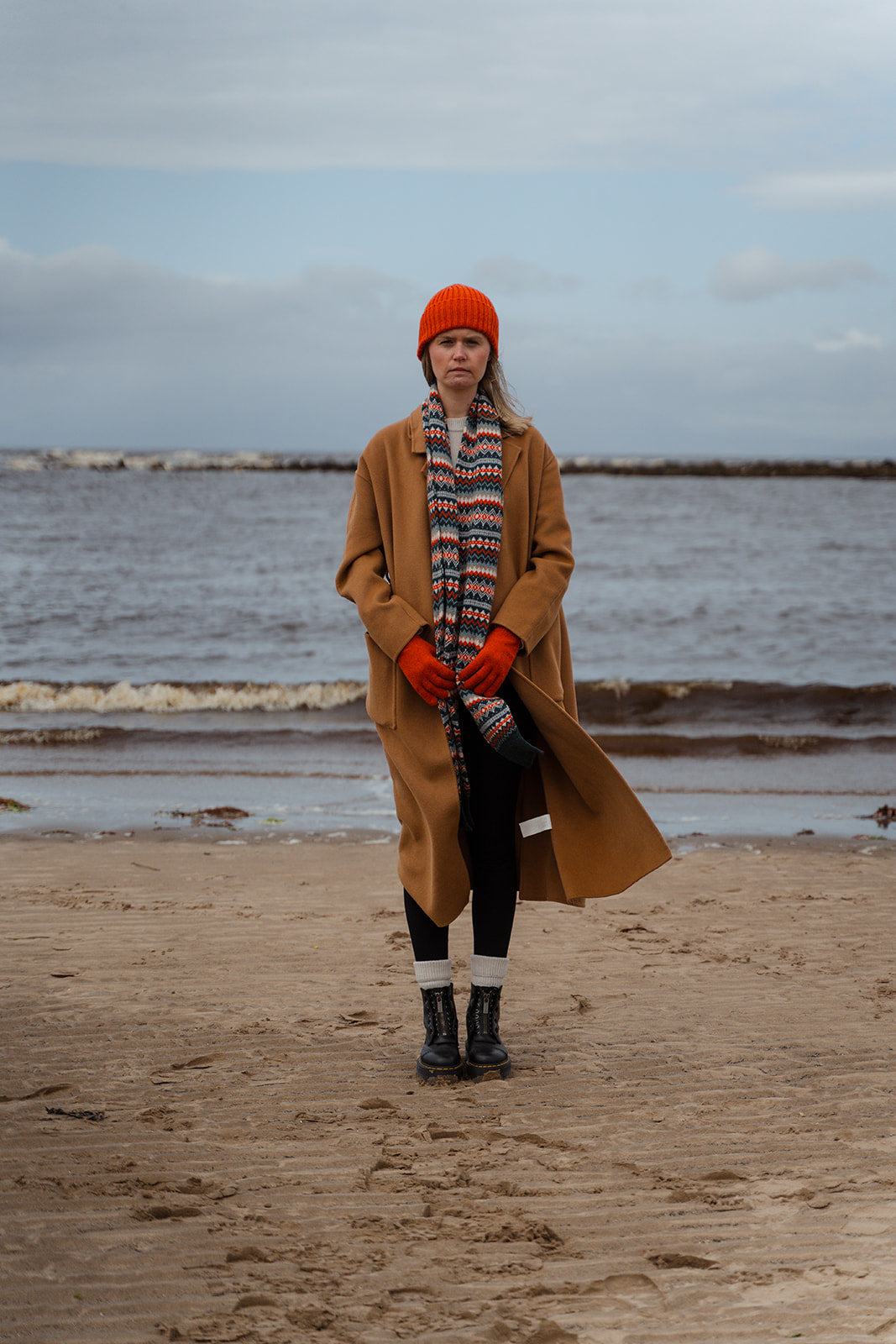 Person wearing a long coat, red hat, and gloves standing on a beach with waves in the background.