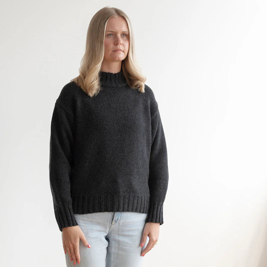 Woman wearing a dark gray sweater and light gray pants against a white background