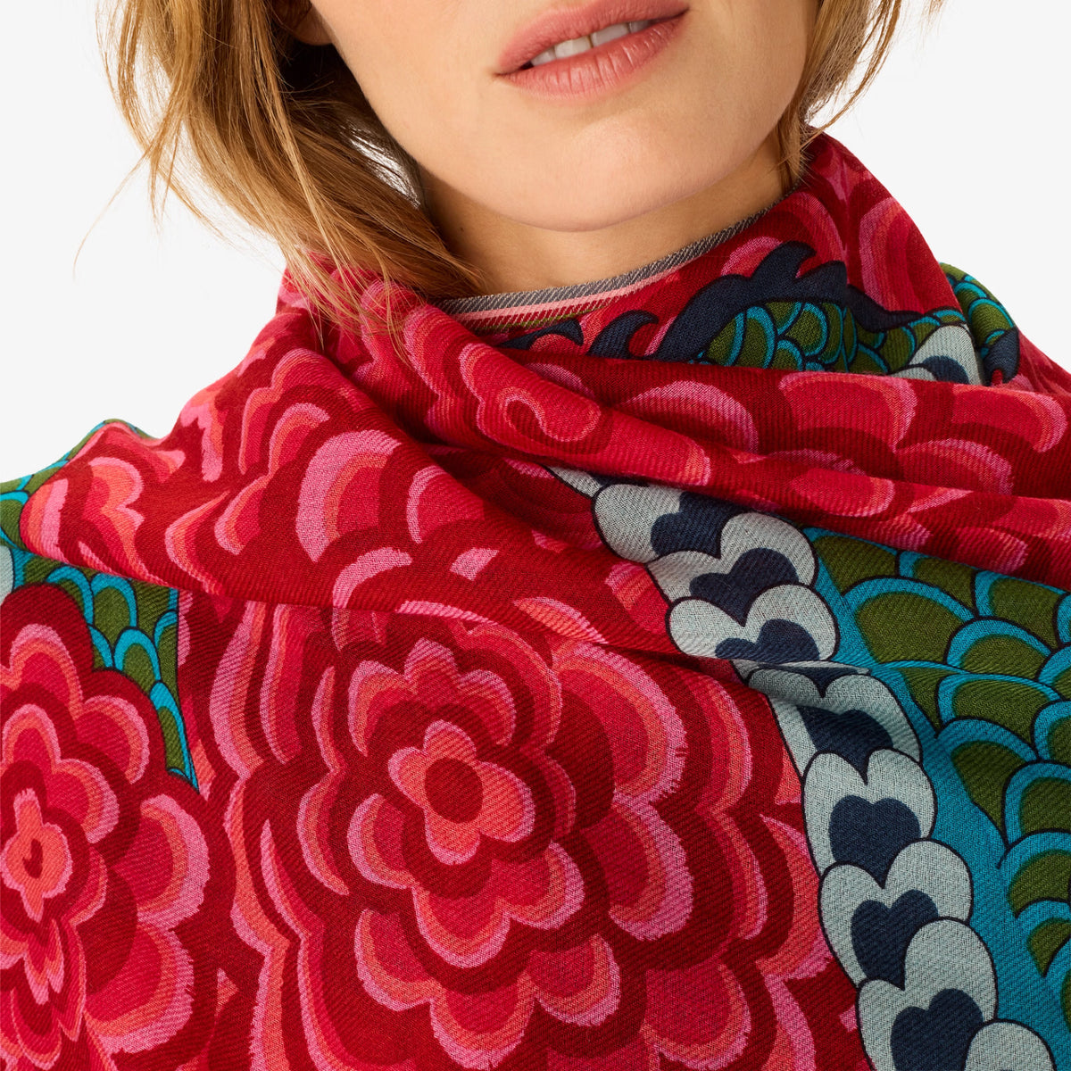 A close-up of a model wearing a vibrant red scarf with a large floral pattern and a contrasting border of teal, green, and gray scalloped design.