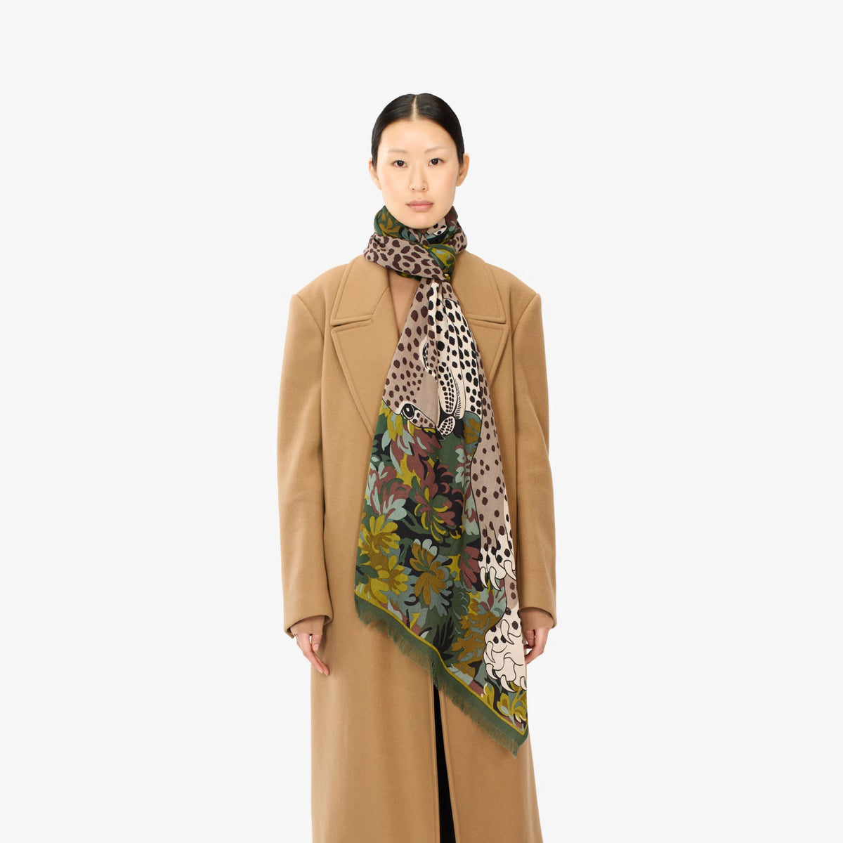 A model wearing a camel-colored coat and a multi-patterned scarf with leopard print, floral designs, and green borders, photographed against a solid white background.