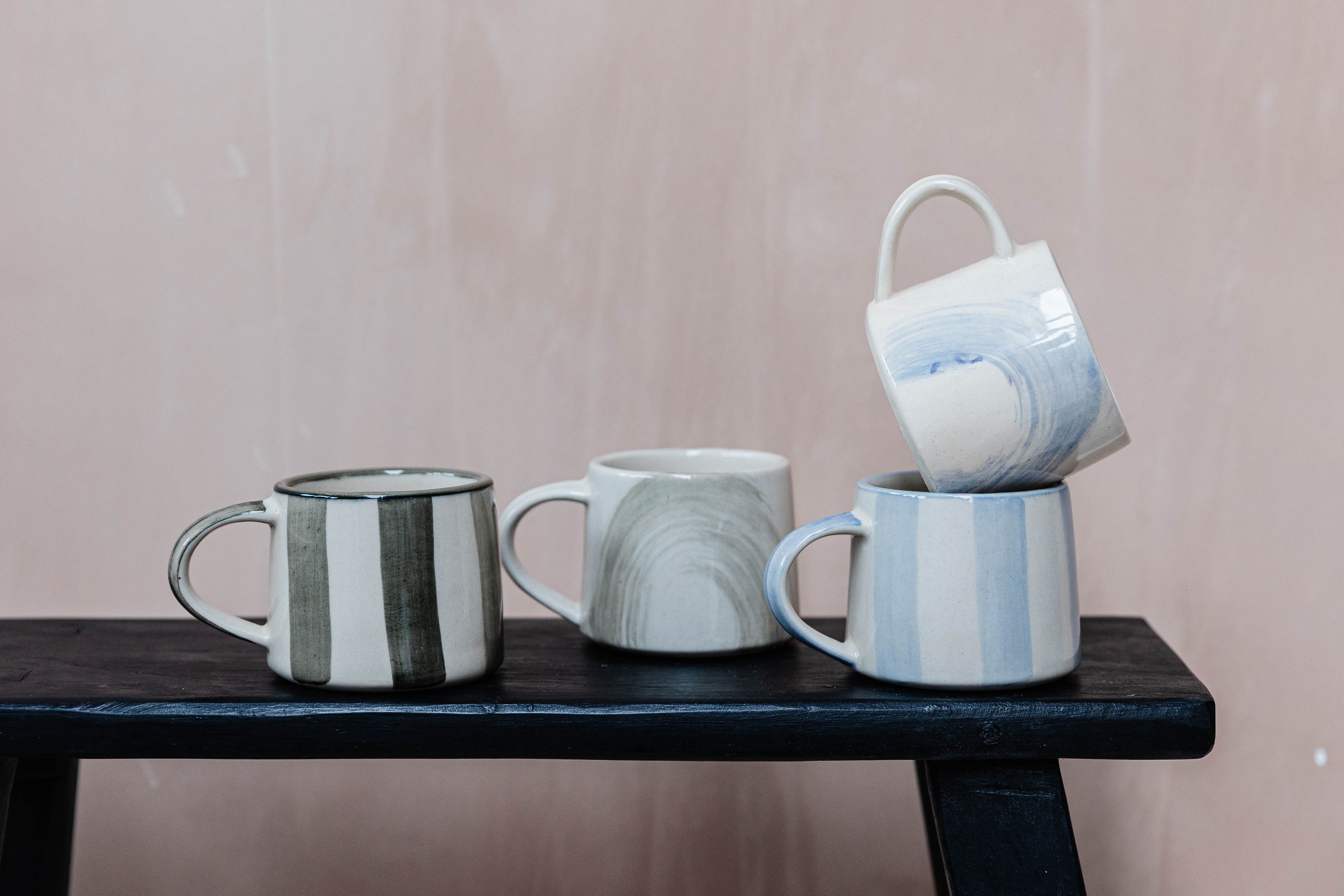 Four hand-painted ceramic mugs in varying stripe and swirl designs.  Mugs are stacked and displayed on a dark wooden surface.