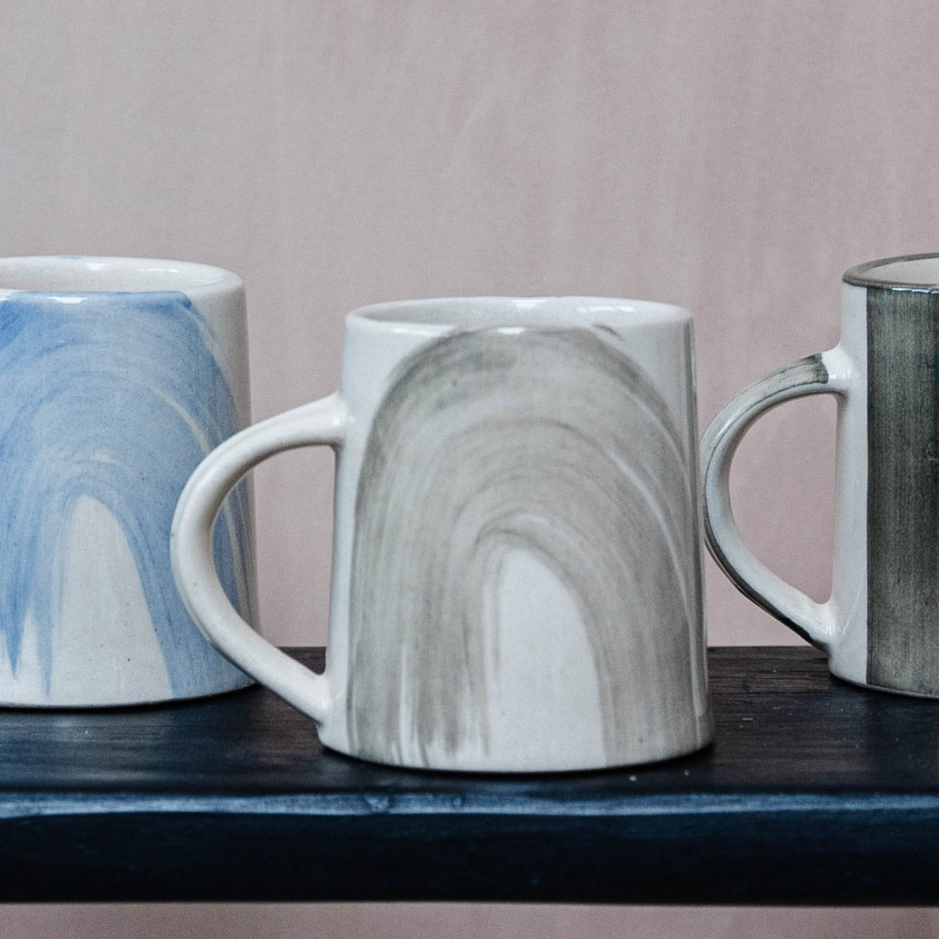 Three ceramic mugs with abstract painted designs; one blue, one grey, and one dark grey.