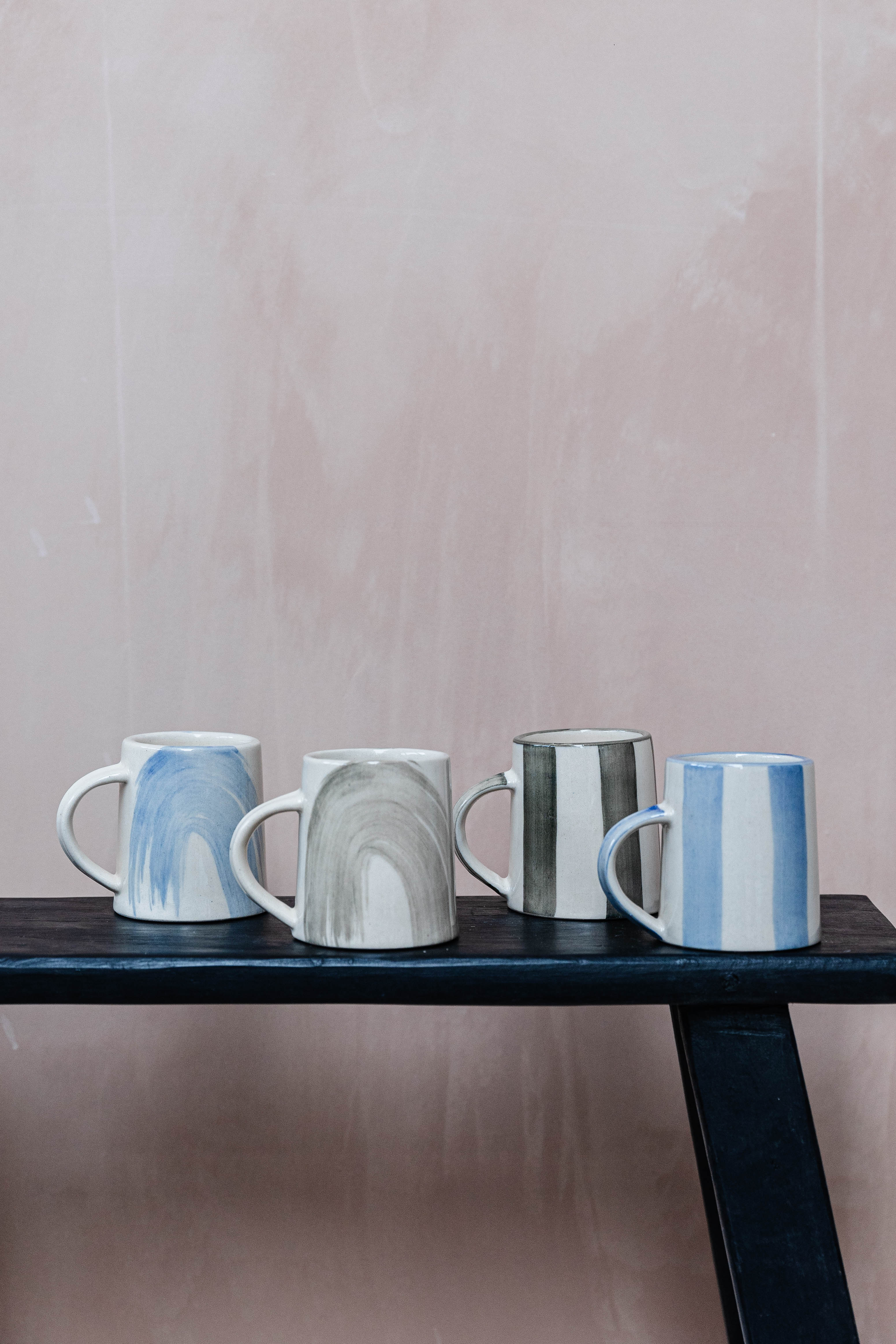 Four ceramic mugs with unique designs: two with abstract blue and grey brushstrokes, two with vertical stripes in blue and