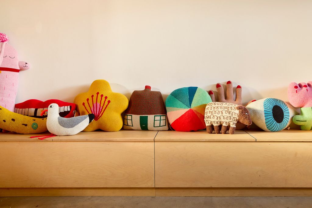 A row of colorful, whimsically shaped plush pillows and soft toys lined up on a light wood bench against a plain beige wall.