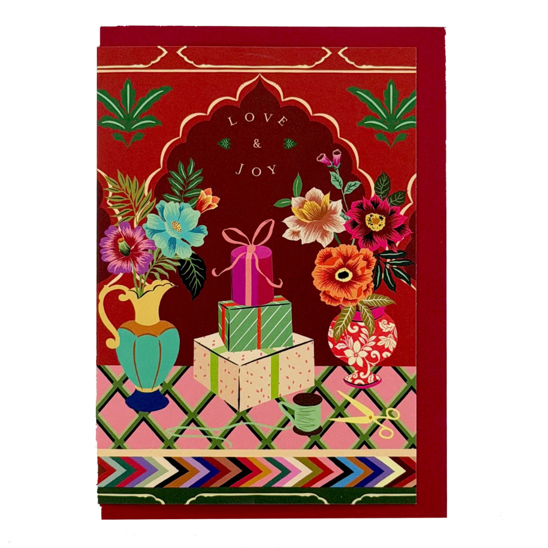 A square greeting card featuring a festive illustration with stacked gift boxes, colorful flowers in vases, and the words 'LOVE & JOY' on a red background with gold decorative borders.