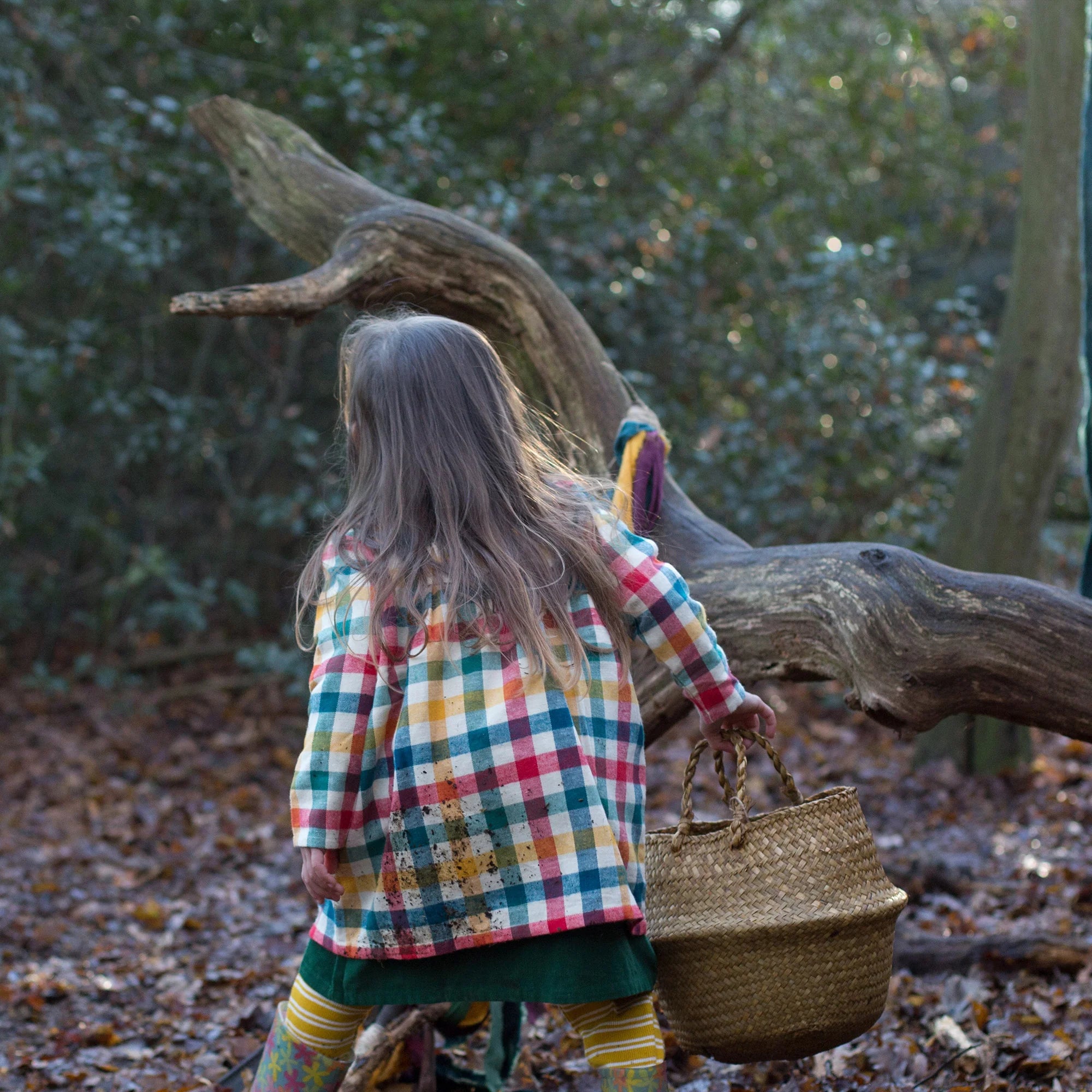 A child, seen from behind, wears a multicolored plaid coat and carries a large, light brown woven basket.