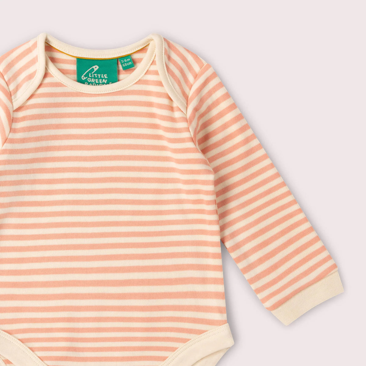 A close-up of a baby long-sleeve bodysuit with horizontal peach and cream stripes, featuring a green 'Little Mermaid' neck label, set against a solid pale pink background.