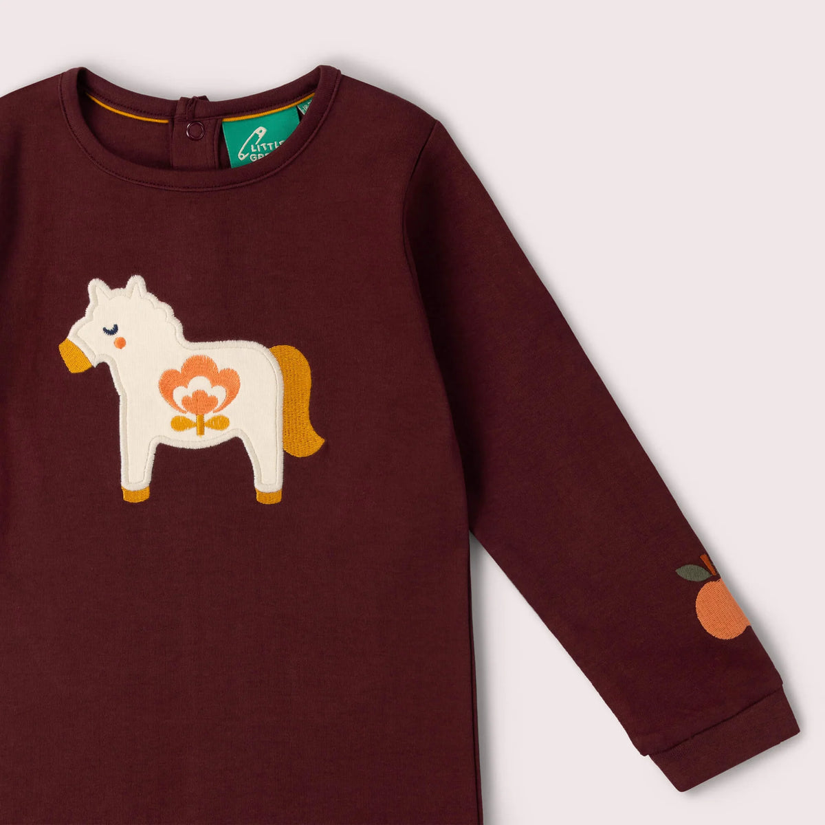 A close-up of a long-sleeve children's top in deep burgundy, featuring a white wooden horse applique with orange and yellow details on the chest and an orange patch on the sleeve.