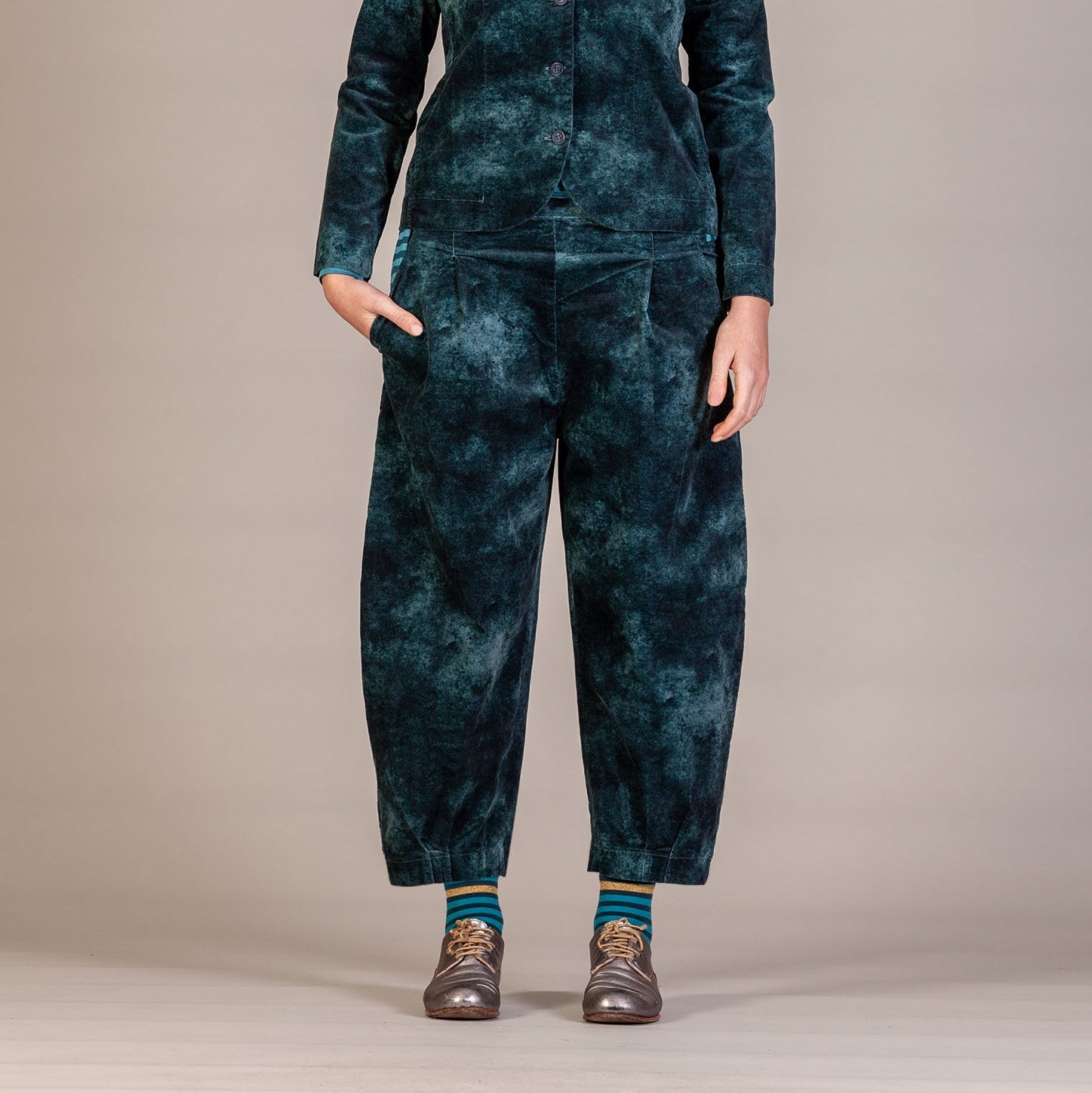 A model stands against a neutral studio backdrop wearing a dark teal and black tie-dye fleece jumpsuit with a stand-up collar and front zipper.