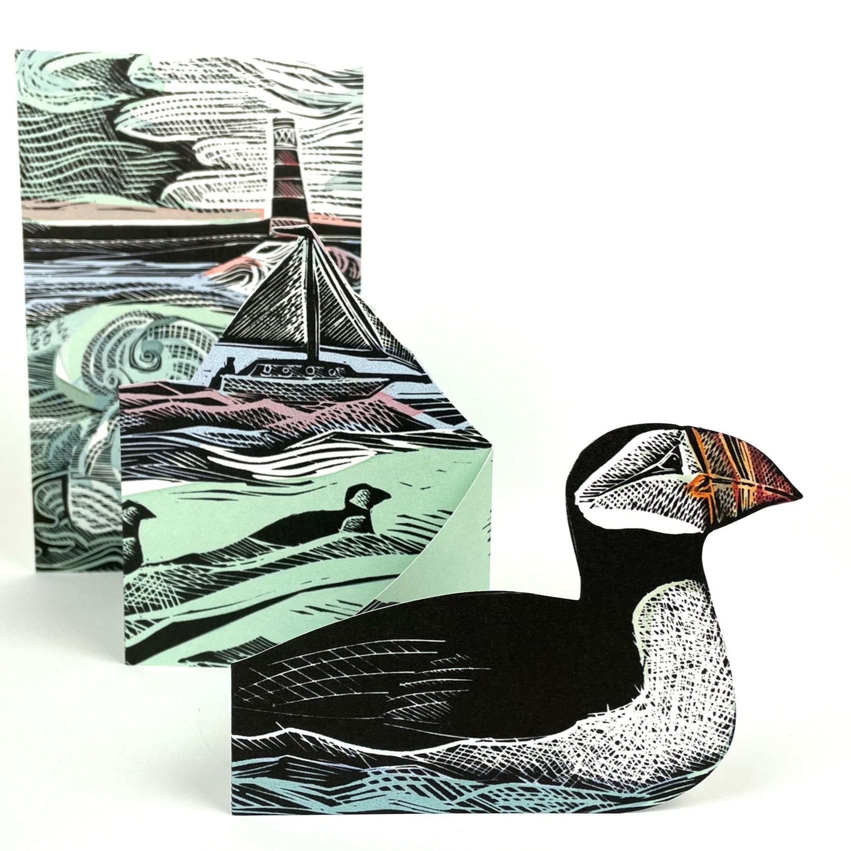 Intricate linocut print of a puffin, shown as a freestanding figure, with a folded card featuring a seascape and sailboat in