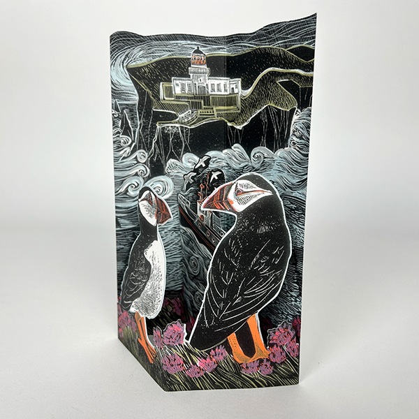 Intricate pop-up card depicting two puffins near a coastal lighthouse, rendered in a detailed, woodcut-style illustration.