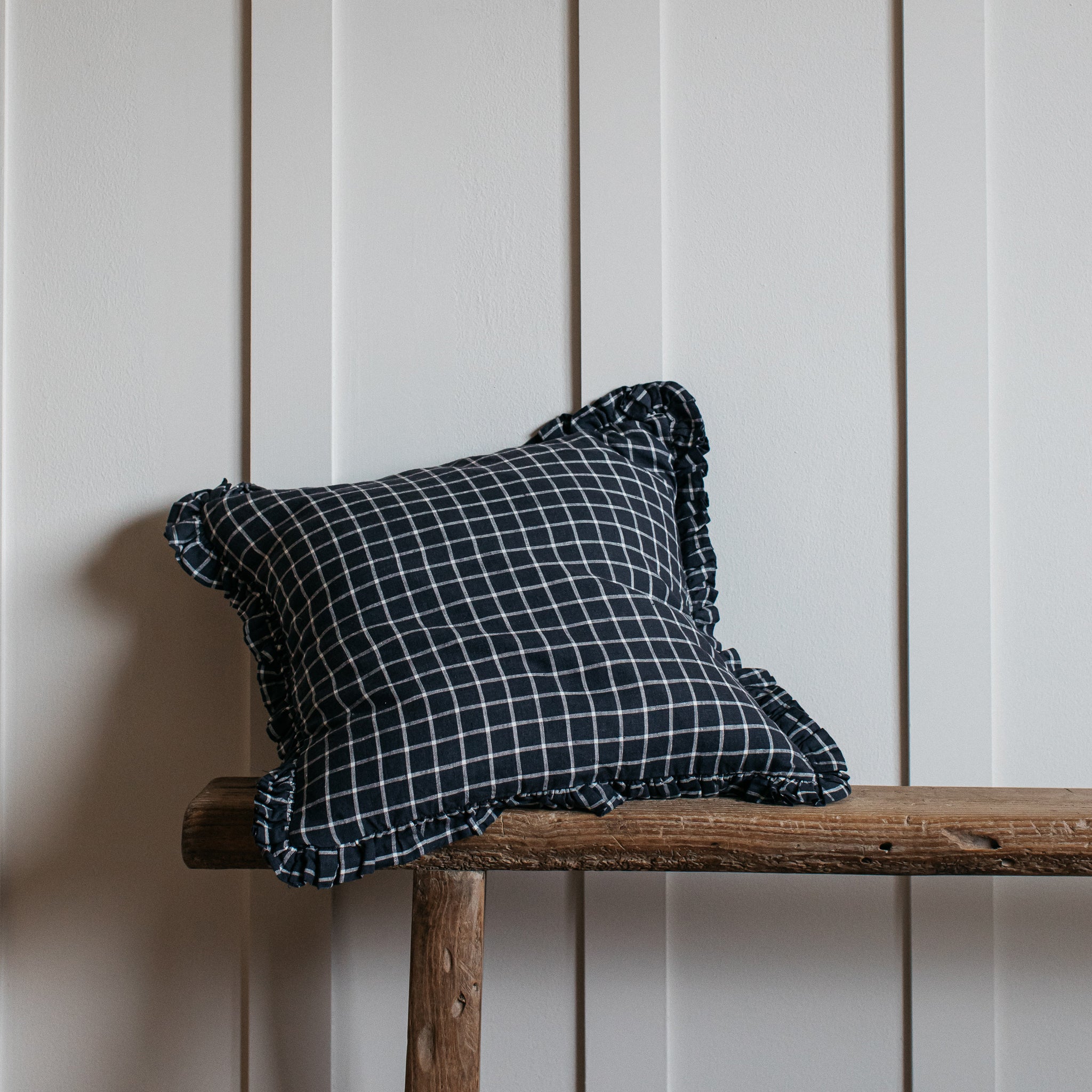 Dark navy blue gingham cushion with ruffled edges, resting on a rustic wooden bench.