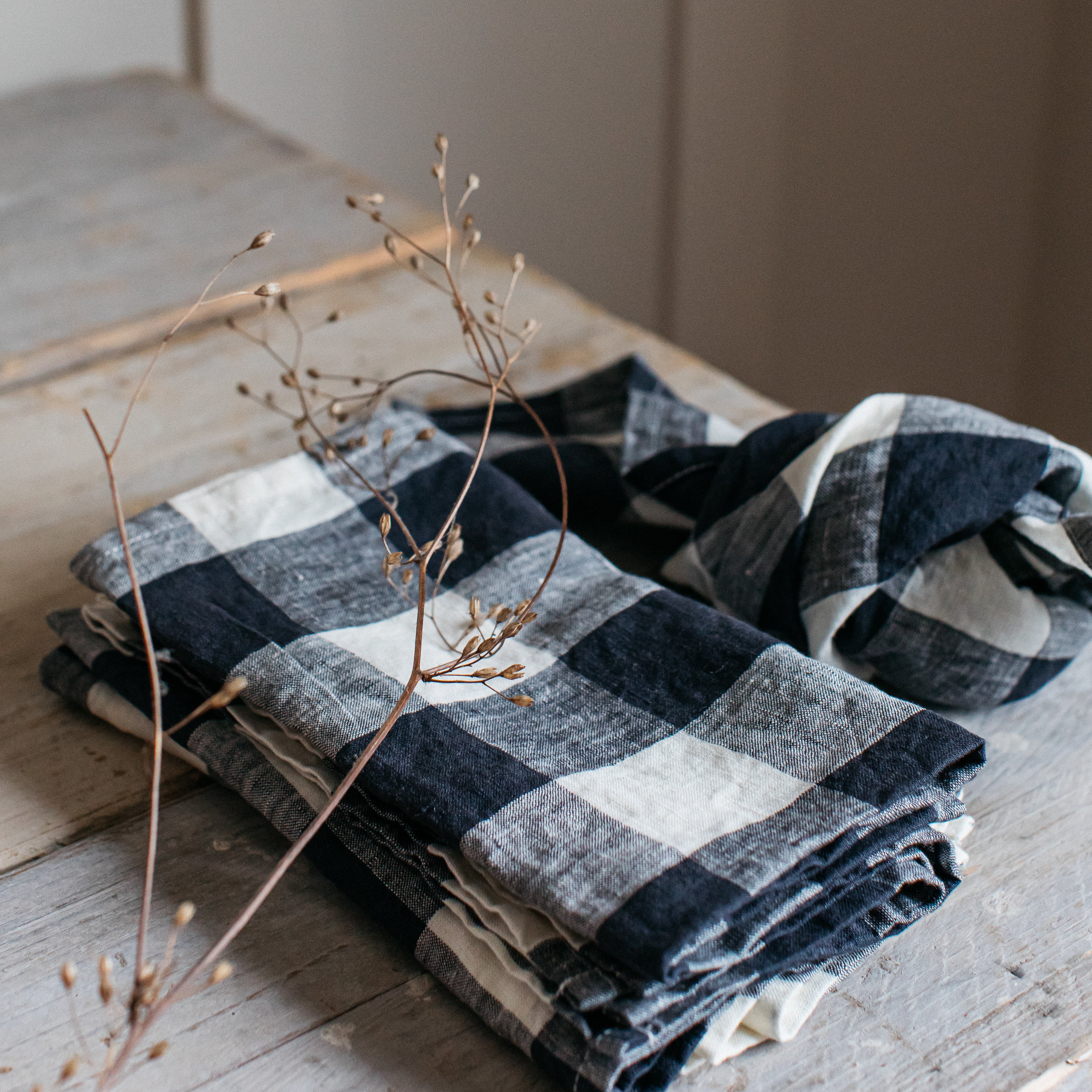 Stack of black and white checkered linen napkins folded on a wooden surface.  A dried plant stem rests on top.