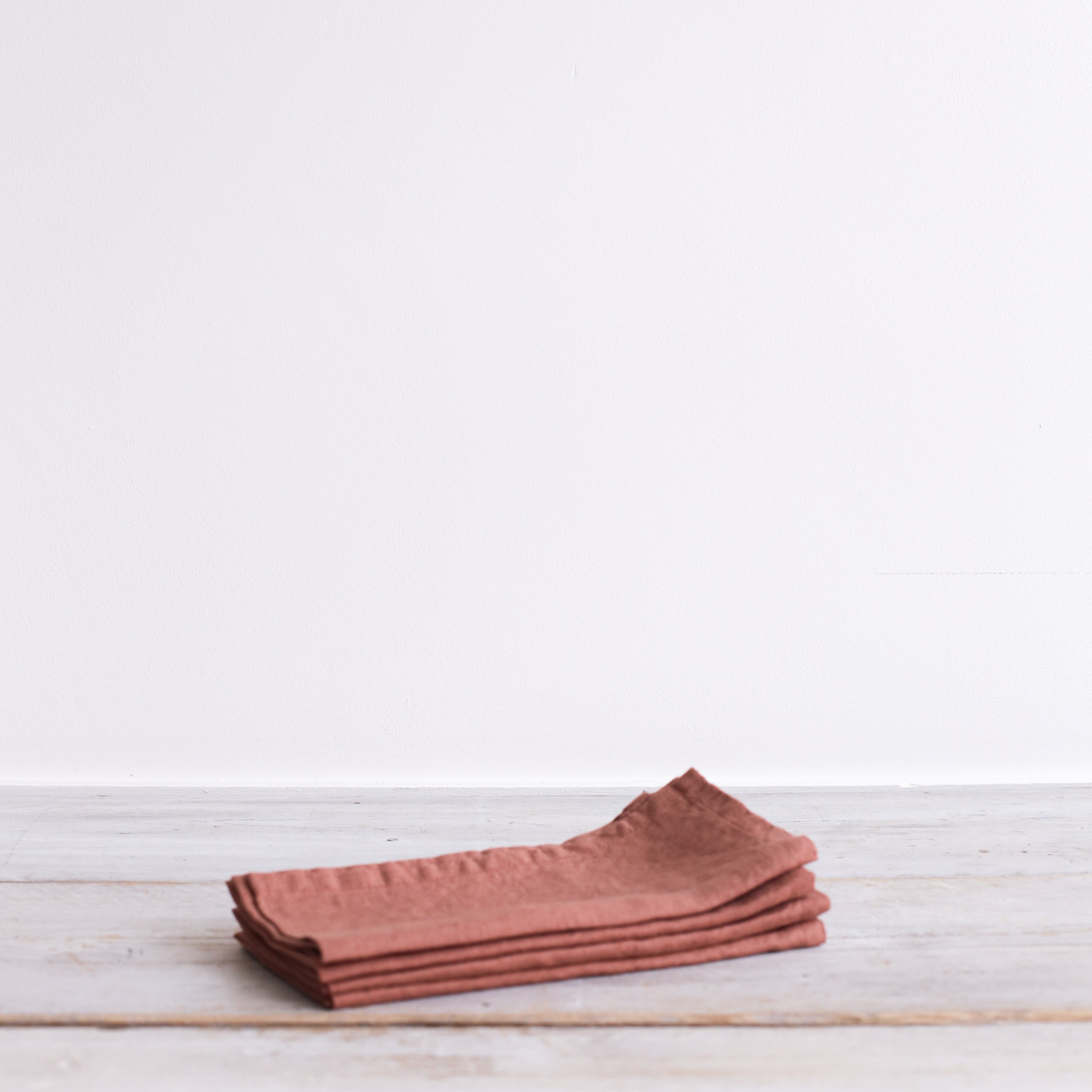 A stack of four terracotta-colored linen napkins on a white wooden surface.