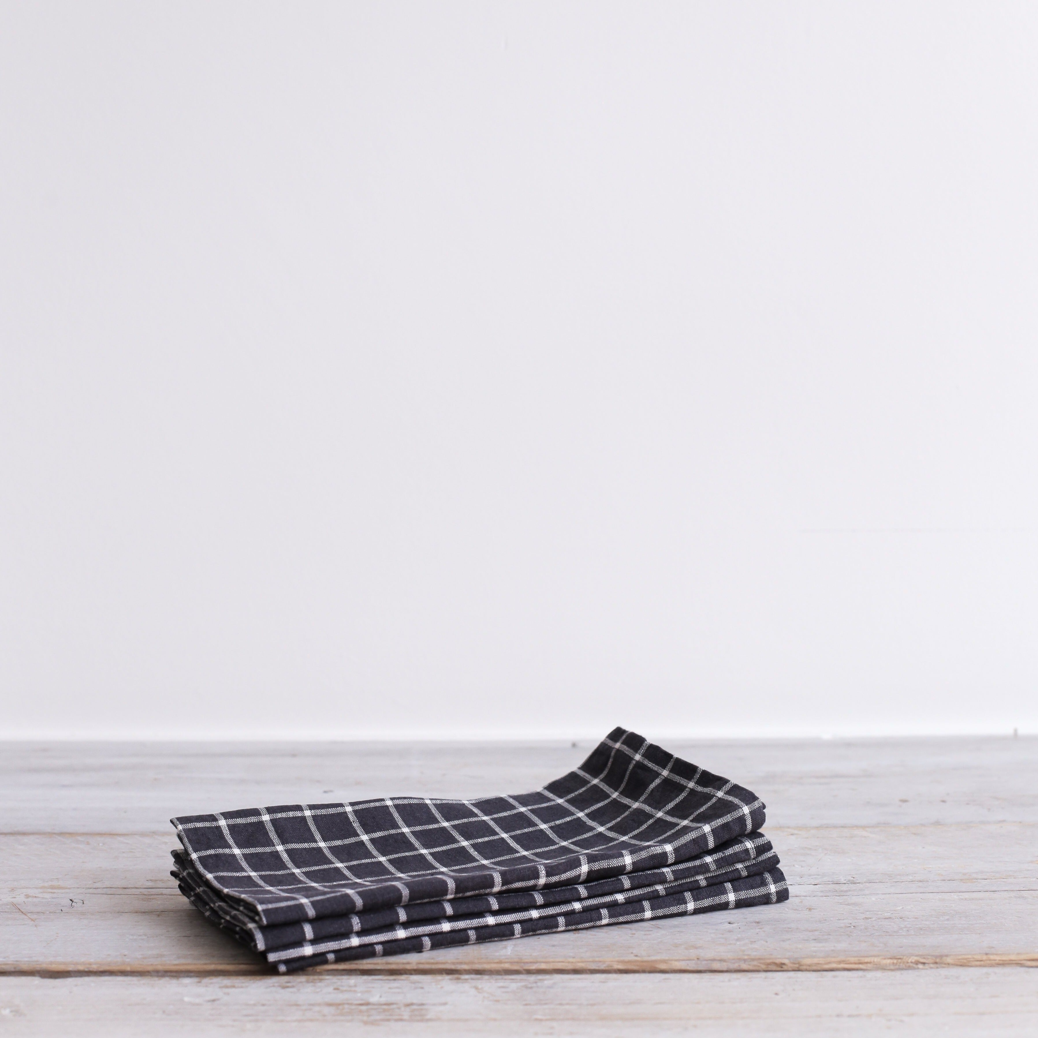 Stack of dark gray linen tea towels with a white grid pattern, folded neatly on a light wood surface.