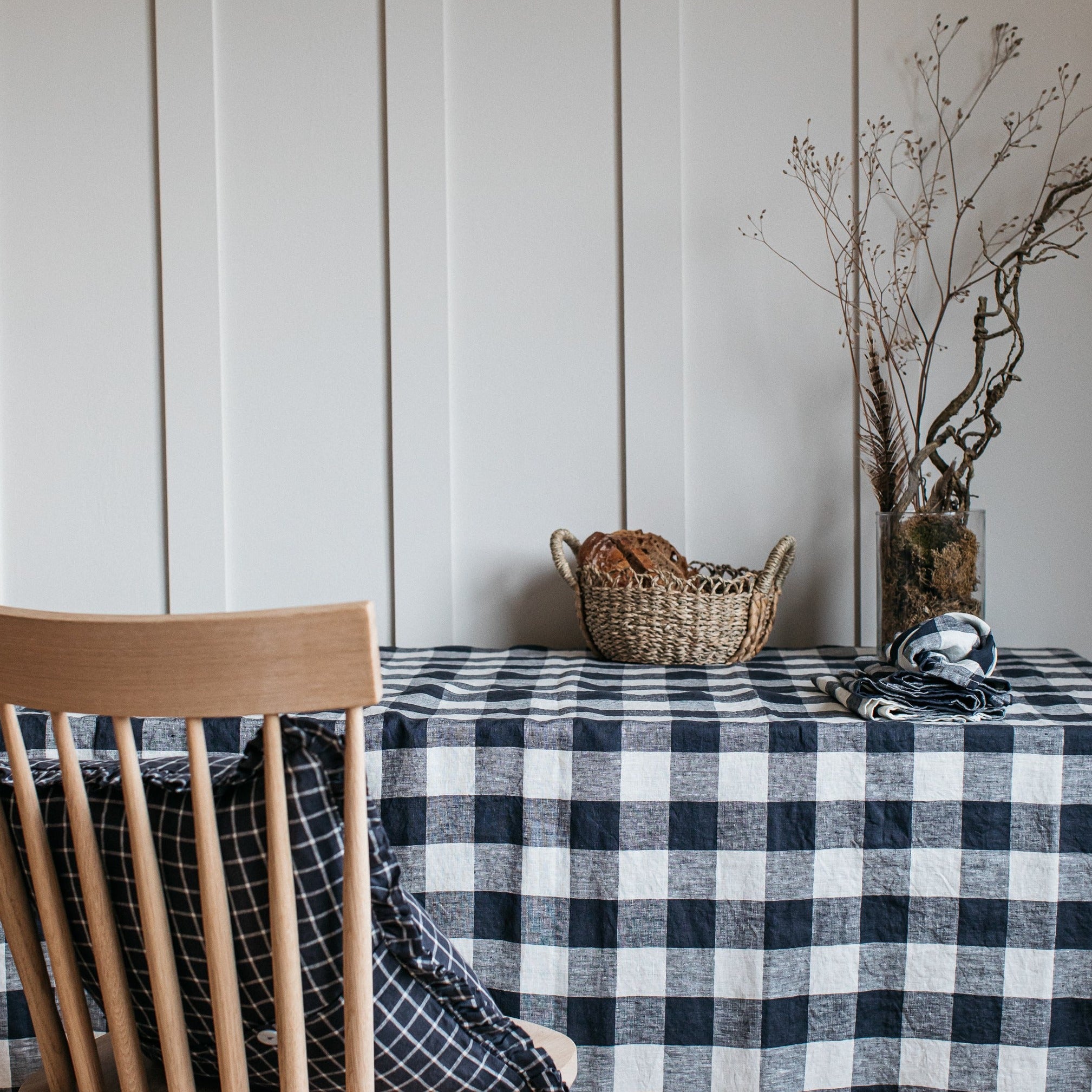 Black and white gingham linen tablecloth draped over a table, with a woven basket of bread and a wooden chair with a