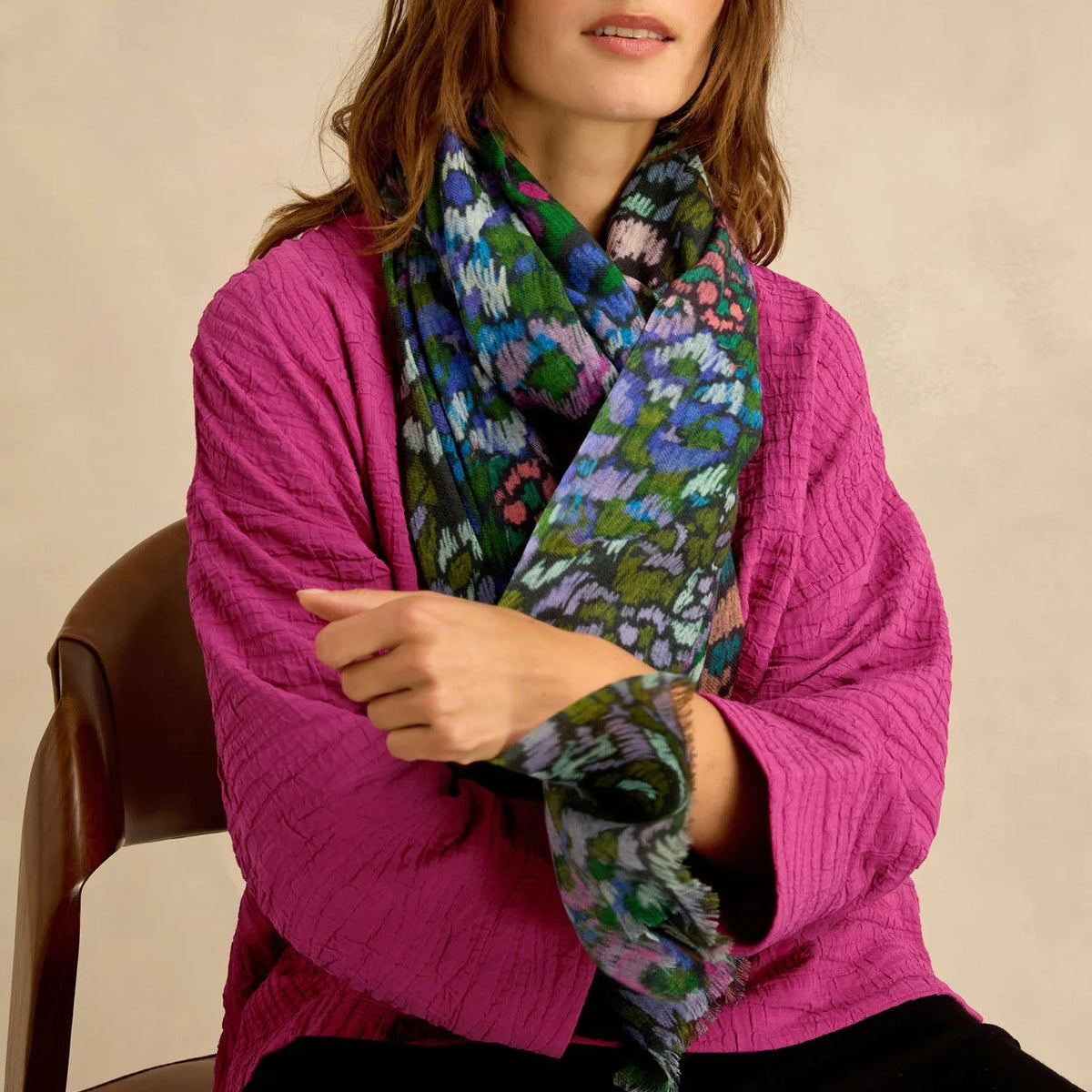 A woman wearing a vibrant magenta quilted cardigan and a green, blue, and purple floral patterned scarf, seated on a wooden chair against a neutral beige background.