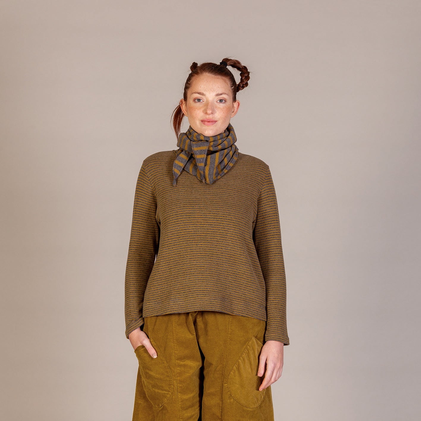 A model stands against a neutral studio backdrop wearing a brown knit turtleneck sweater, wide-leg mustard yellow pants, a plaid neck gaiter, and brown lace-up boots.