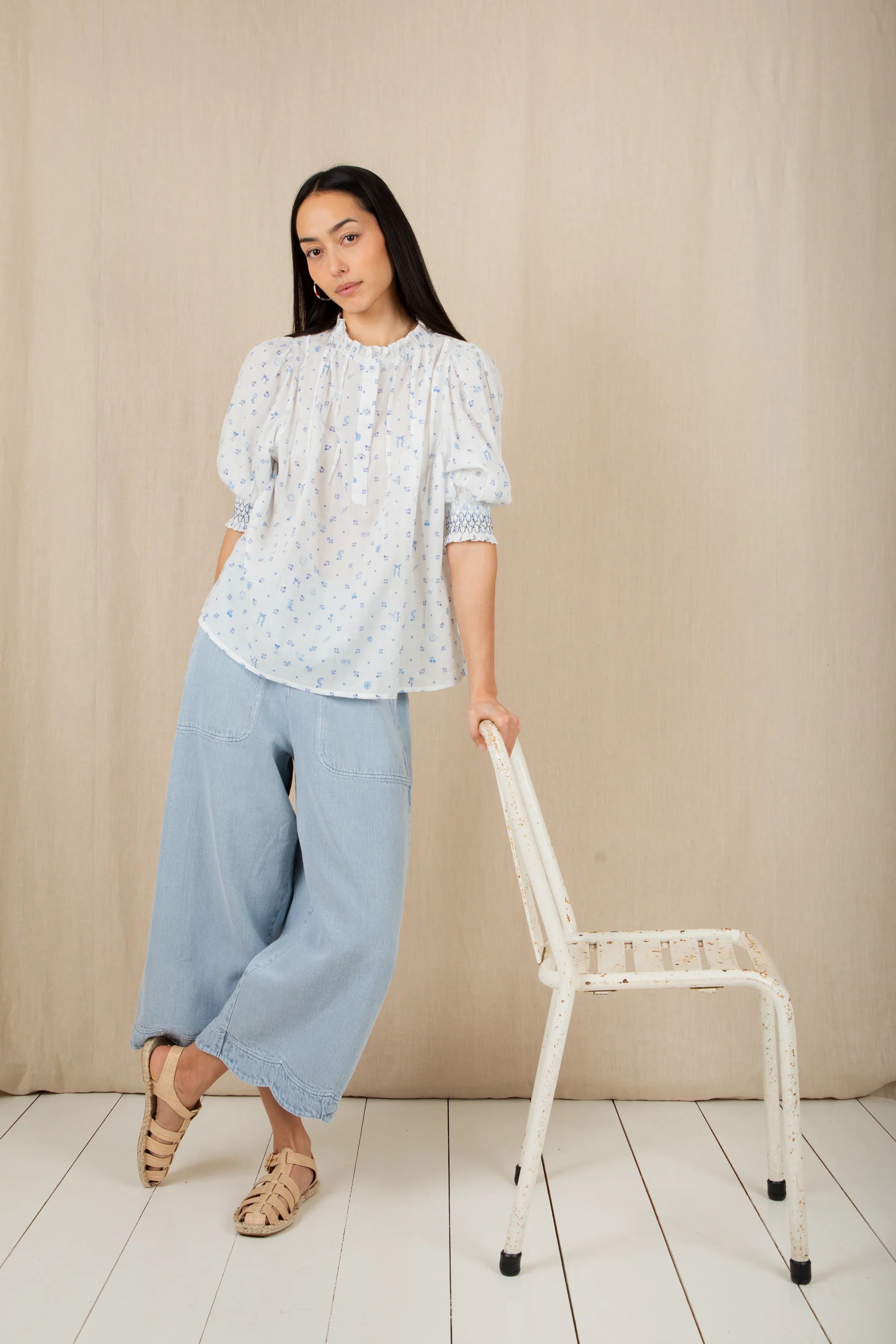 Woman in a light blue floral blouse and wide-leg pants standing next to a white chair against a beige wall.