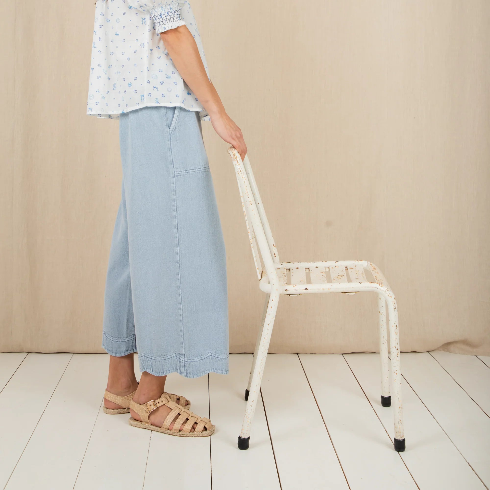 Woman in a light-colored outfit standing next to a white chair against a beige wall.