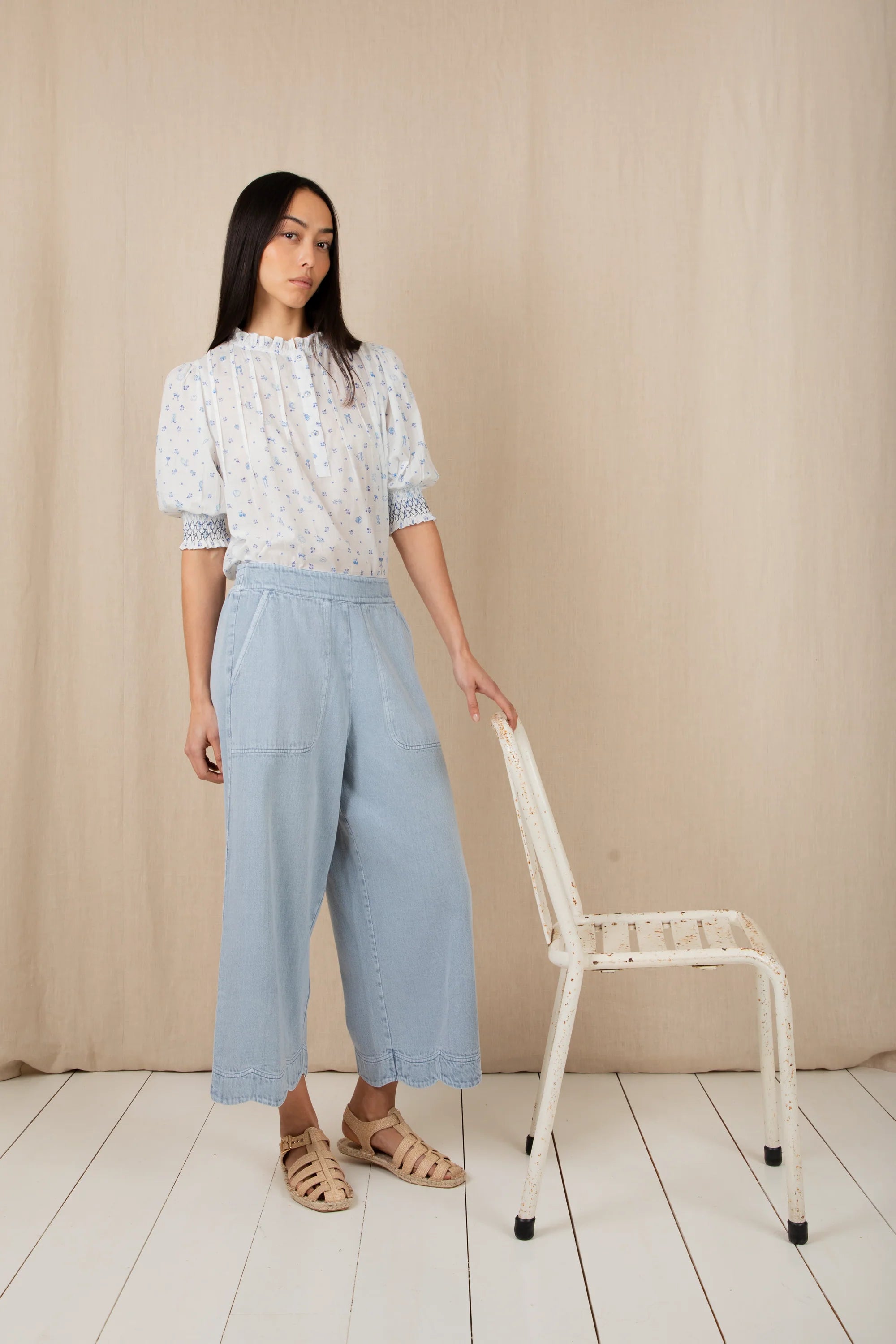 Woman in a floral blouse and light blue pants standing next to a white chair against a beige wall.