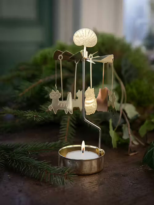 Decorative candle holder with a small lamp and hanging figures on a wooden surface with greenery in the background.