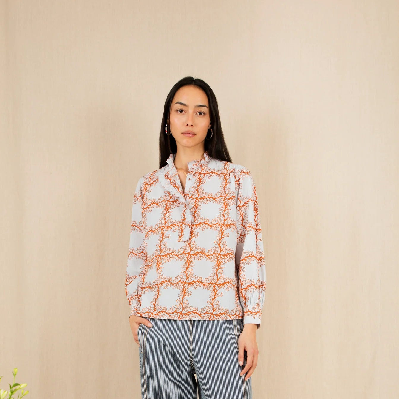 Woman wearing a patterned shirt and jeans standing against a beige wall with decorative plants.