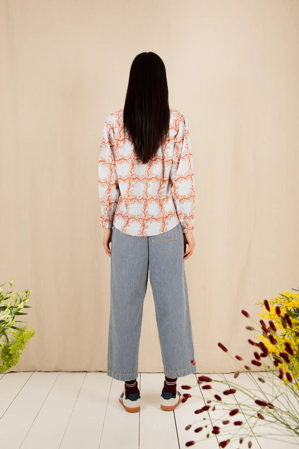 Person wearing a patterned shirt and jeans standing against a beige wall with plants on either side.