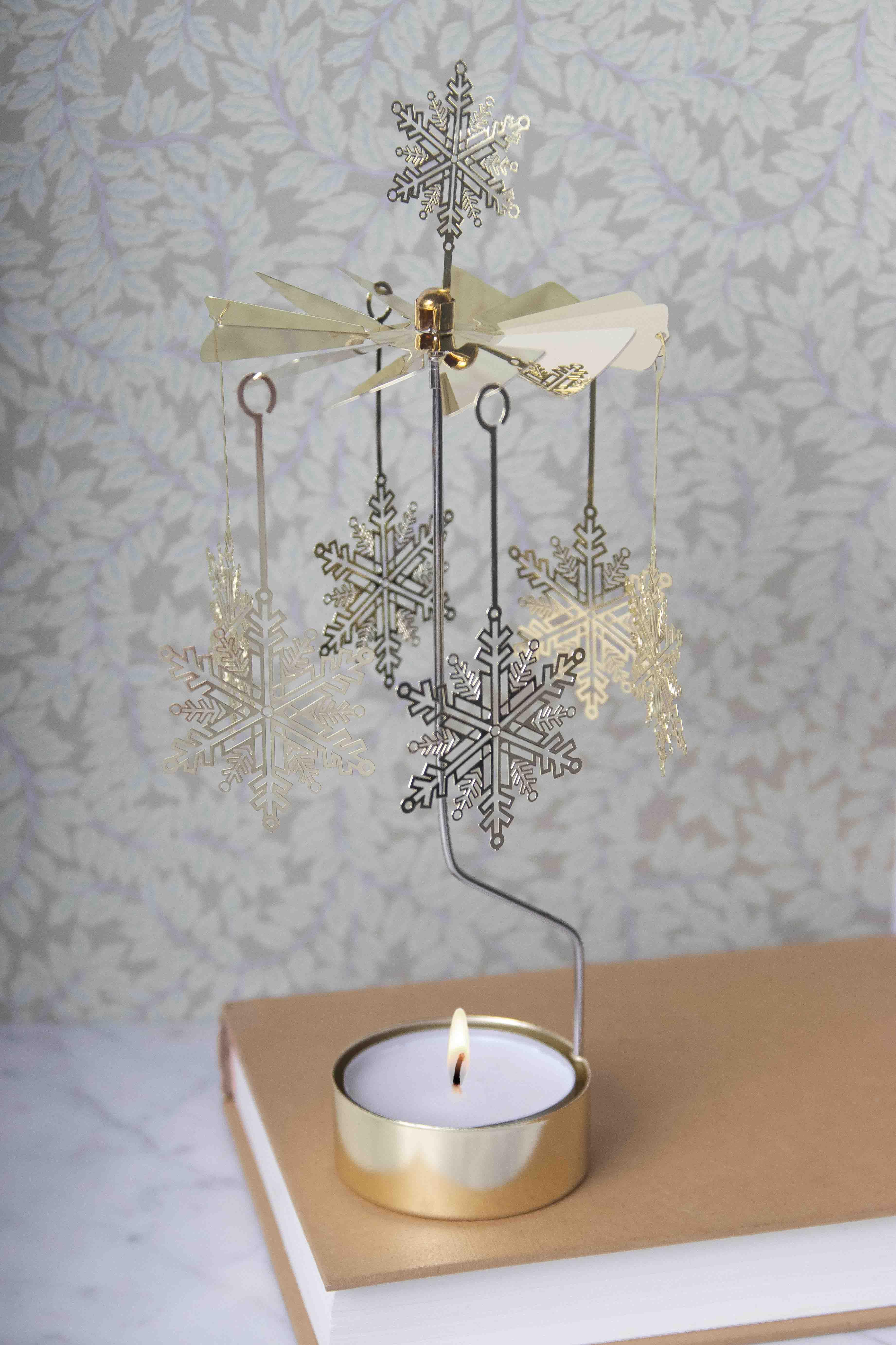 A decorative snowflake wind spinner with hanging snowflakes and a gold-toned metal stand, shown lit with a small candle in a gold holder on top of a book against a patterned wallpaper background.