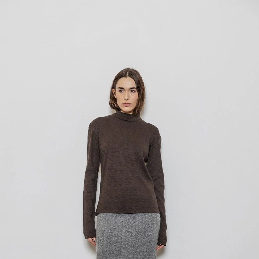 Woman wearing a brown turtleneck sweater and gray skirt with black knee-high boots against a white wall.