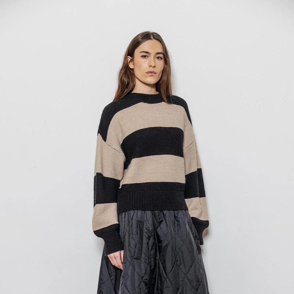 Woman wearing a striped sweater and quilted pants against a white wall.