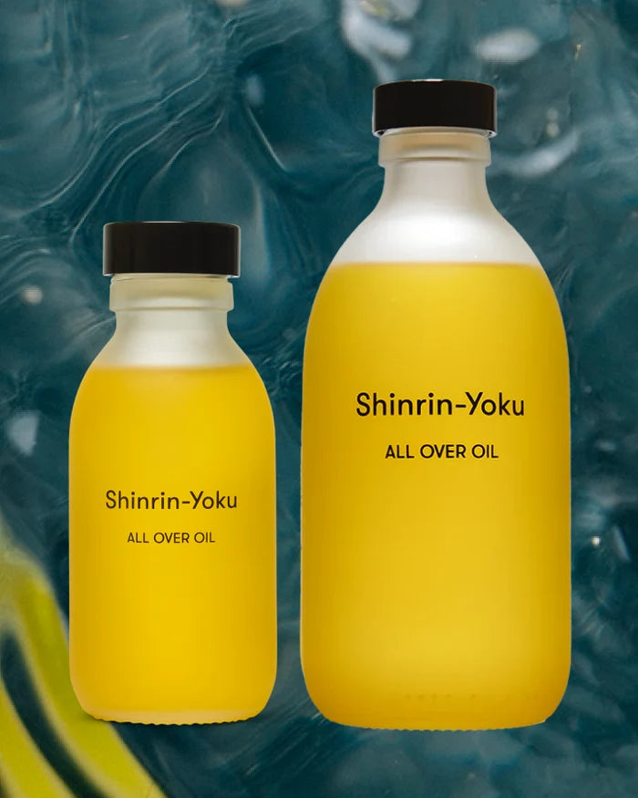 Two frosted glass bottles of Shinrin-Yoku All Over Oil, one small and one large, filled with yellow oil and set against a dark blue marbled background.