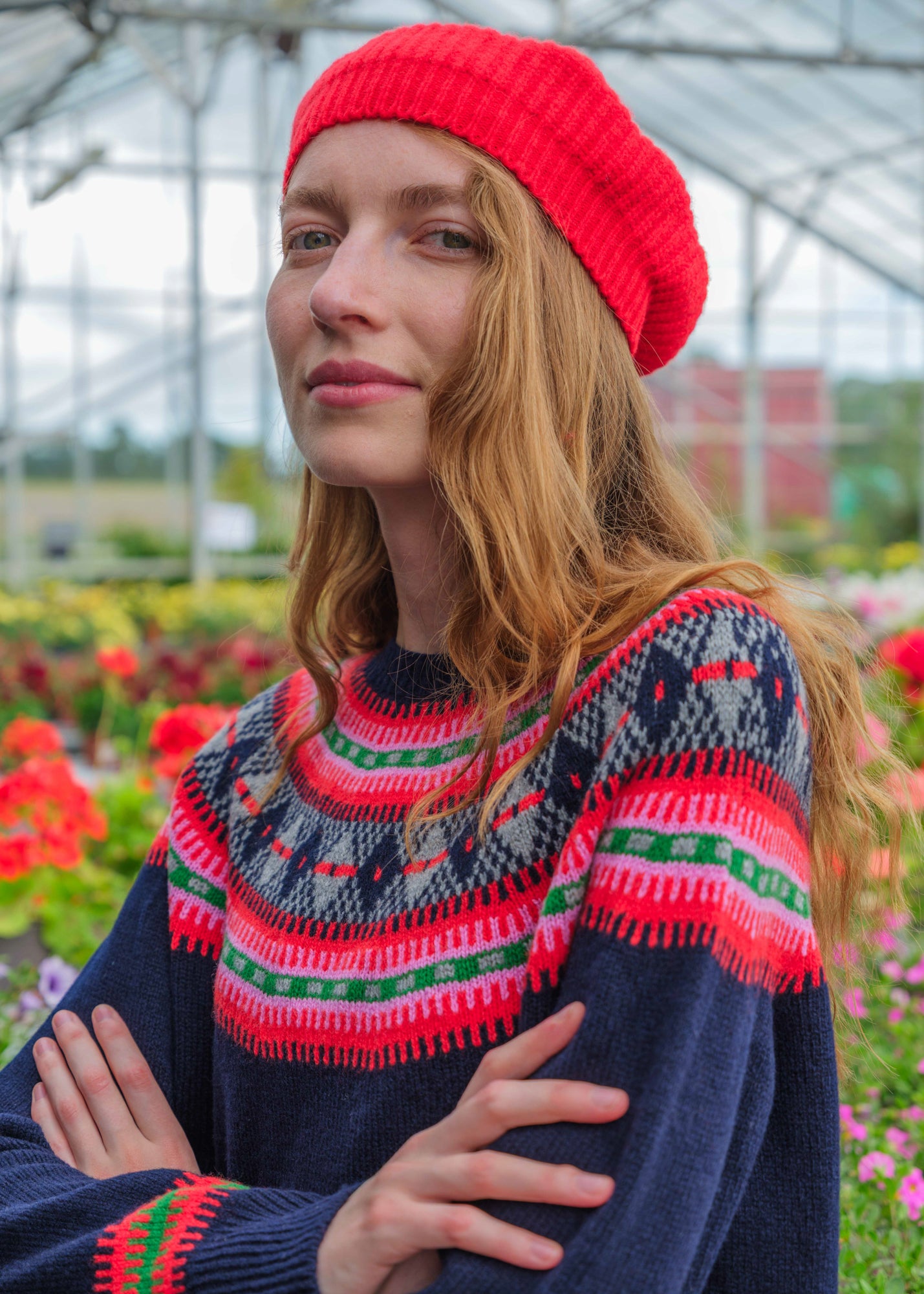 A woman wearing a bright red knit beanie and a navy Fair Isle sweater with red, pink, and green patterns, standing in a flower garden greenhouse.