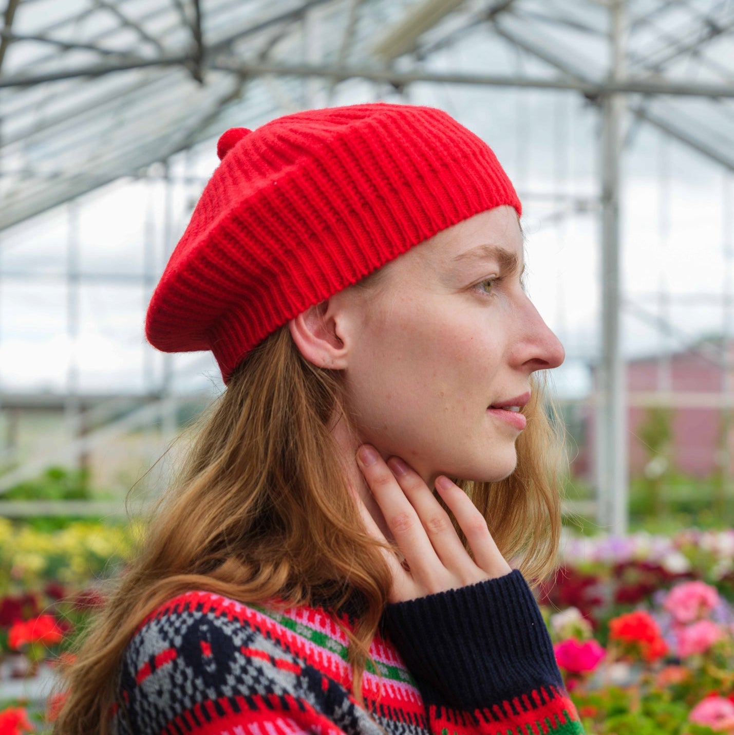 A woman in a bright red knit beret and a dark navy sweater with red, green, and gray Nordic-style patterns stands in a flower garden greenhouse.