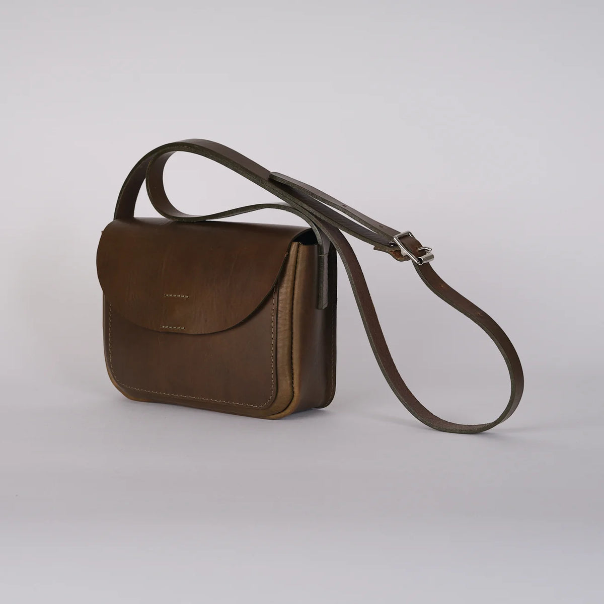 A dark brown leather crossbody bag with a flap front pocket and a matching adjustable shoulder strap, photographed on a clean light gray studio background.