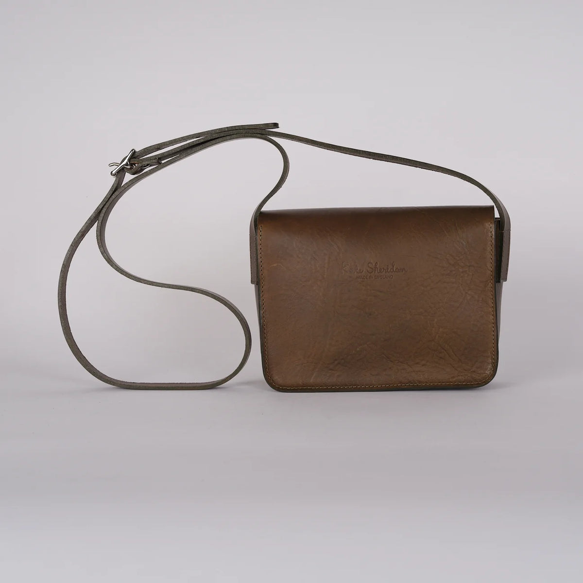 A brown leather crossbody bag with a thin dark gray strap, shown against a neutral light gray background.