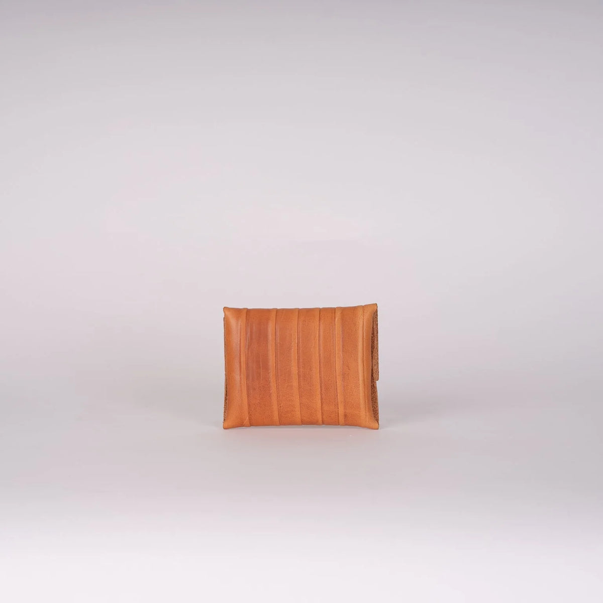 A small rectangular clutch or envelope-style handbag in warm tan leather with vertical stitched paneling, centered on a light gray studio background.