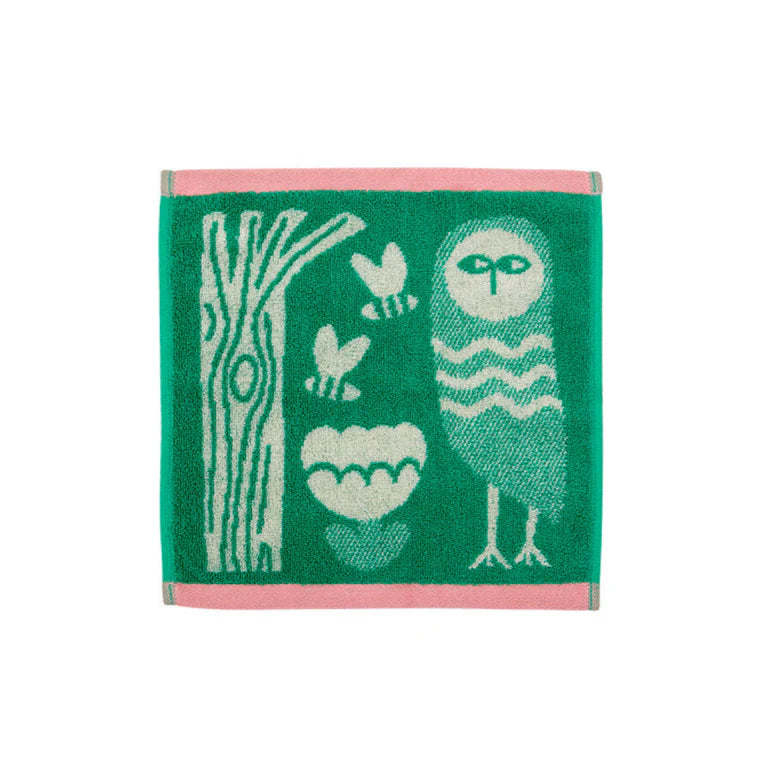 A green towel by Donna Wilson featuring a white owl, tree, and flowers, bordered in pink.
