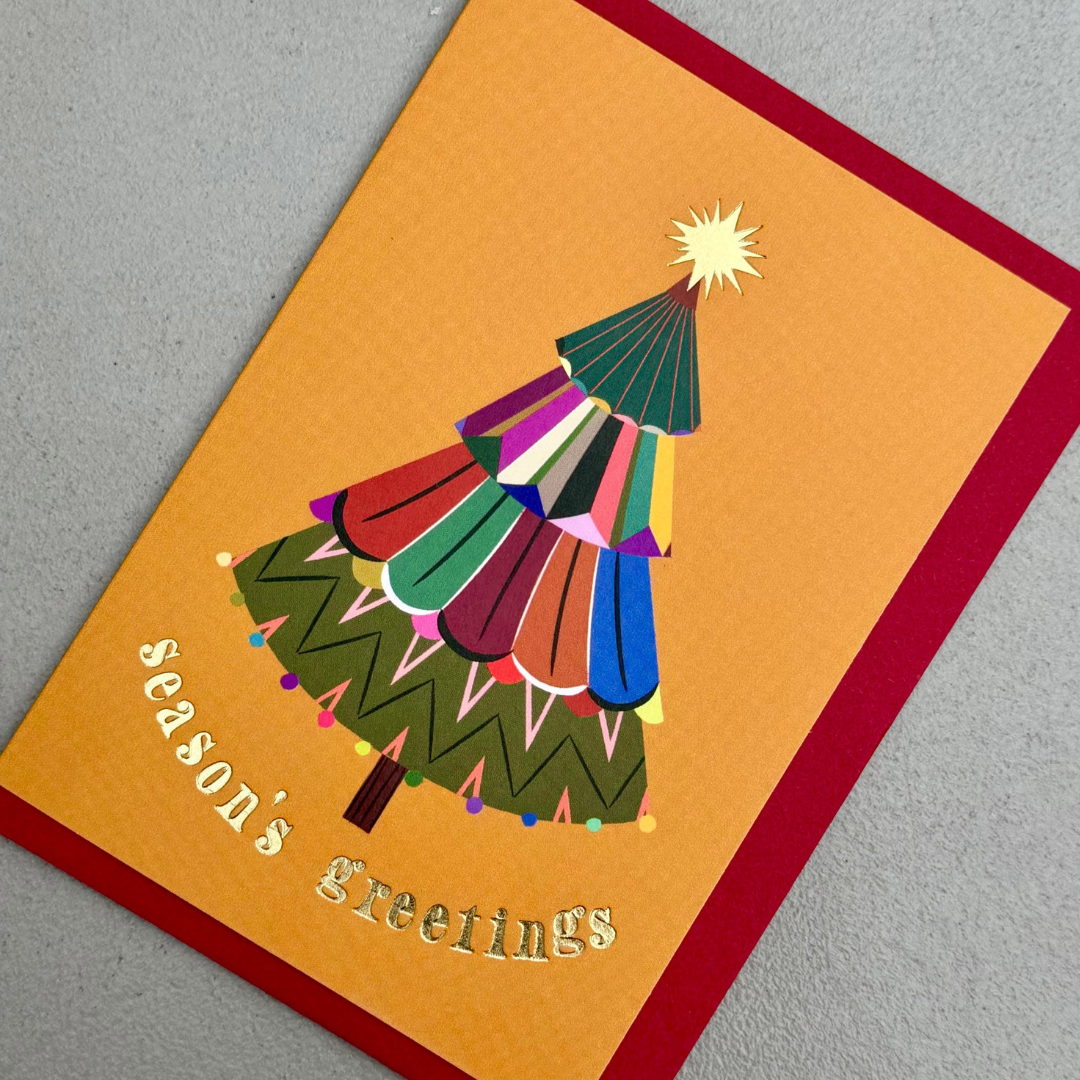 A square Christmas greeting card with a bright orange face, a red border, and a stylized multicolored Christmas tree. The words 'Season's greetings' appear in gold at the lower left, and the card is photographed on a light gray textured surface.