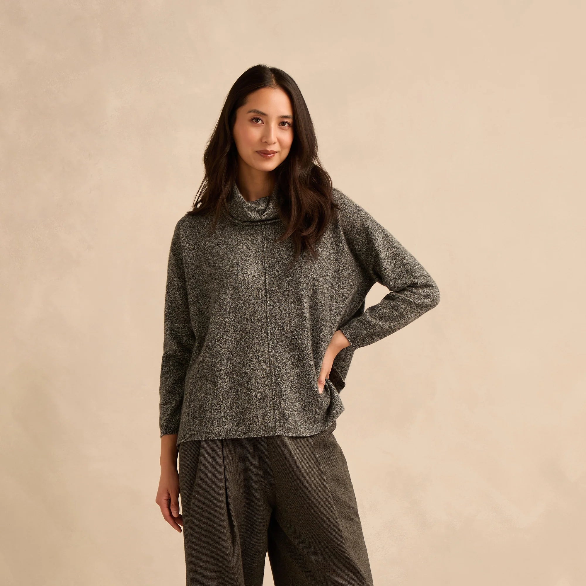 A woman modeling a dark gray textured knit mock-neck sweater and dark olive green wide-leg trousers, paired with brown suede mules, against a warm beige studio backdrop.