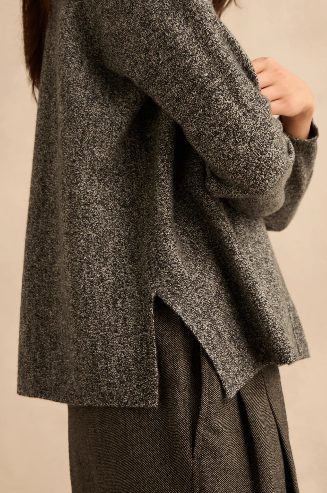 A close-up of a person wearing a dark gray and black heathered knit cardigan with a side slit, paired with matching dark trousers, against a plain beige background.